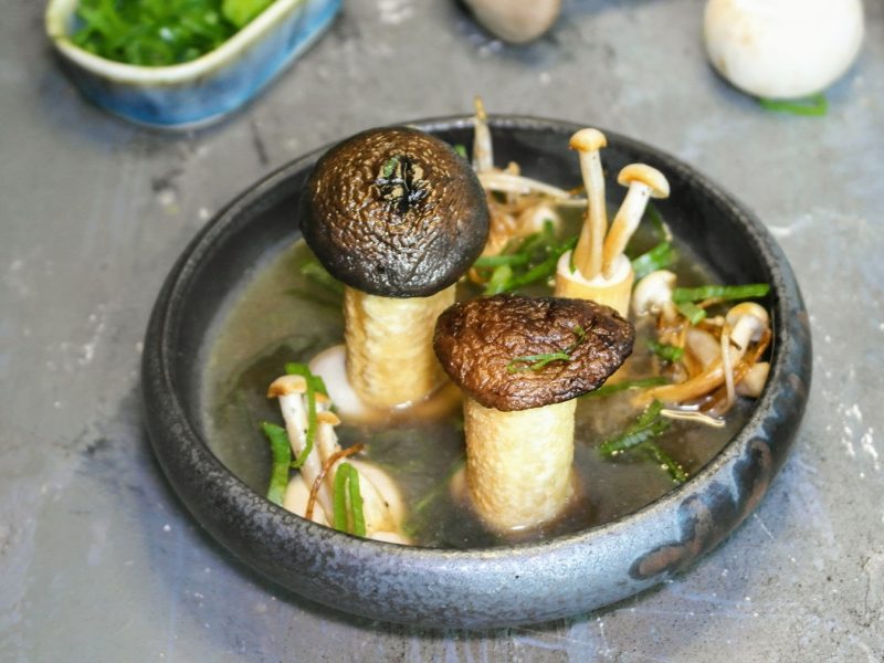 Eine graue Schale mit der Pilzsuppe auf steinernem Untergrund von schräg oben fotografiert. Dahinter Frühlingslauch und ungeschnittene Pilze zu erkennen.