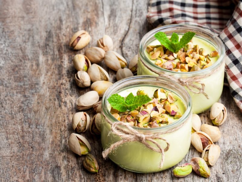 Draufsicht, schräg: Zwei Gläser mit selbst gemachter Pistazien-Mousse, die auf einem Untergrund aus Holz stehen. Drum herum liegen frische Pistazienkerne mit und ohne Schale.