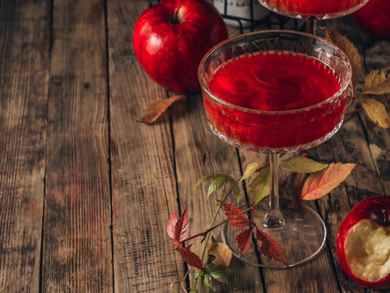 Ein kristallklares Glas mit Poison Apple Cocktail, leuchtende Äpfel und farbige Herbstblätter auf einem rustikalen Holztisch.