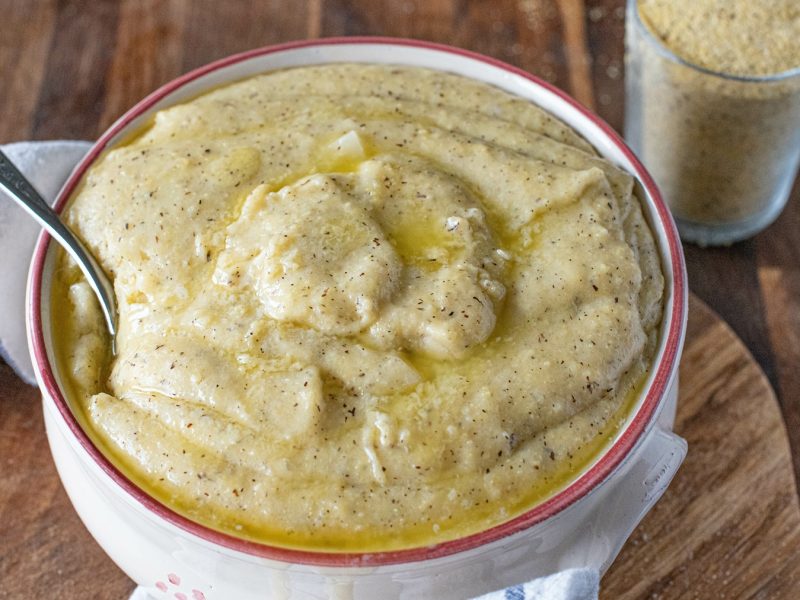 Eine Schüssel mit cremiger Polenta taragna, die mit geschmolzener Butter beträufelt ist, steht auf einem Holzbrett neben einem Glas Polentagrieß.