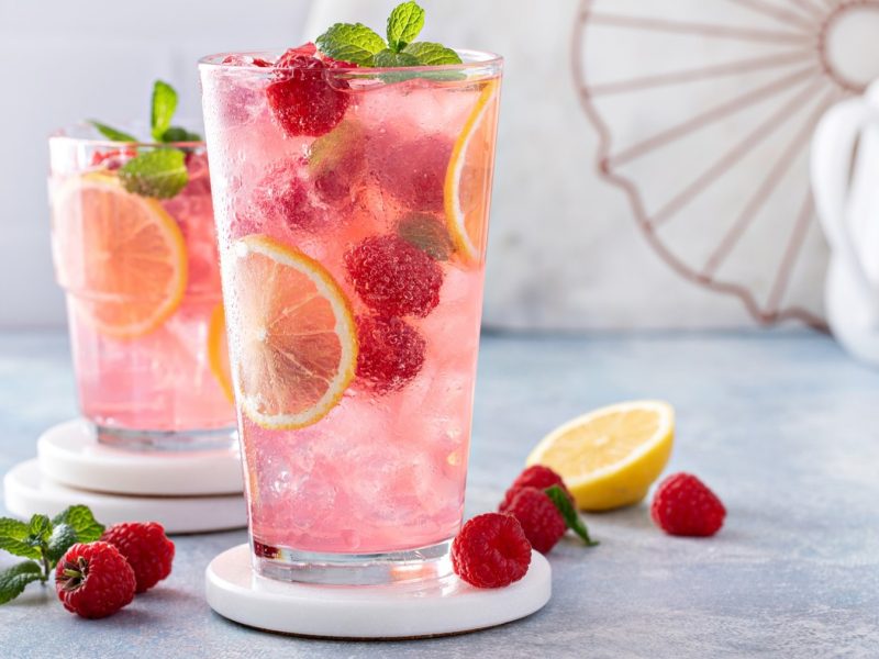 Nahaufnahme eines Glases Raspberry Limoncello mit frischen Himbeeren, Zitronenscheiben und Minzblättern, arrangiert auf einem sommerlichen Tisch.