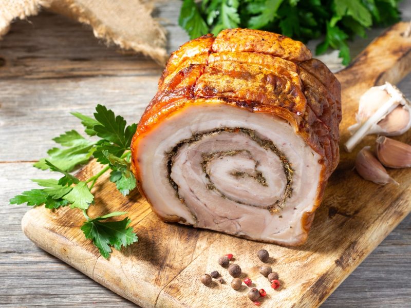 Ein angeschnittener Rollbraten auf einem Holzbrett mit Petersilie und Pfefferkörnern von schräg oben fotografiert.