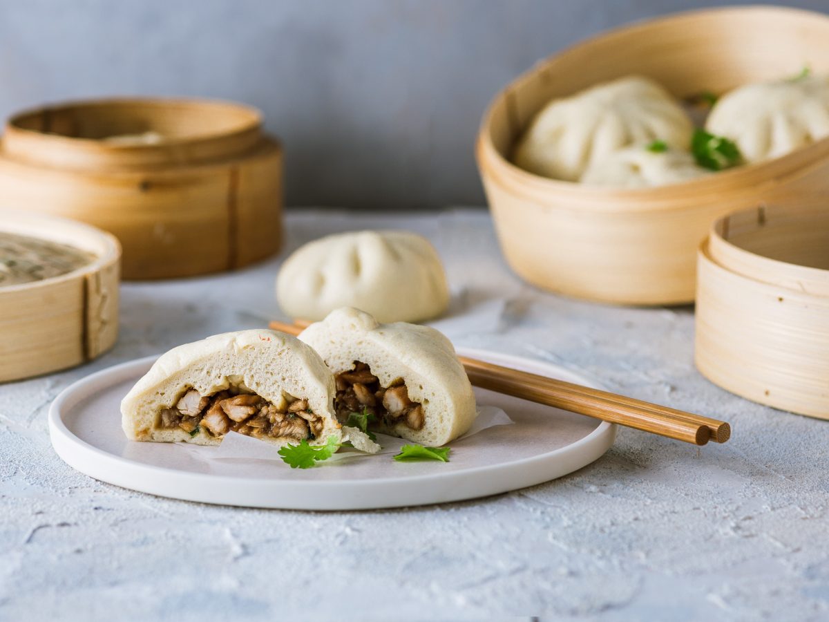 Die Dumplings auf einem hellen Teller und grauem Unter- und Hintergrund mit Dampfkörben und Essstäbchen. Alles von vorne fotografiert.