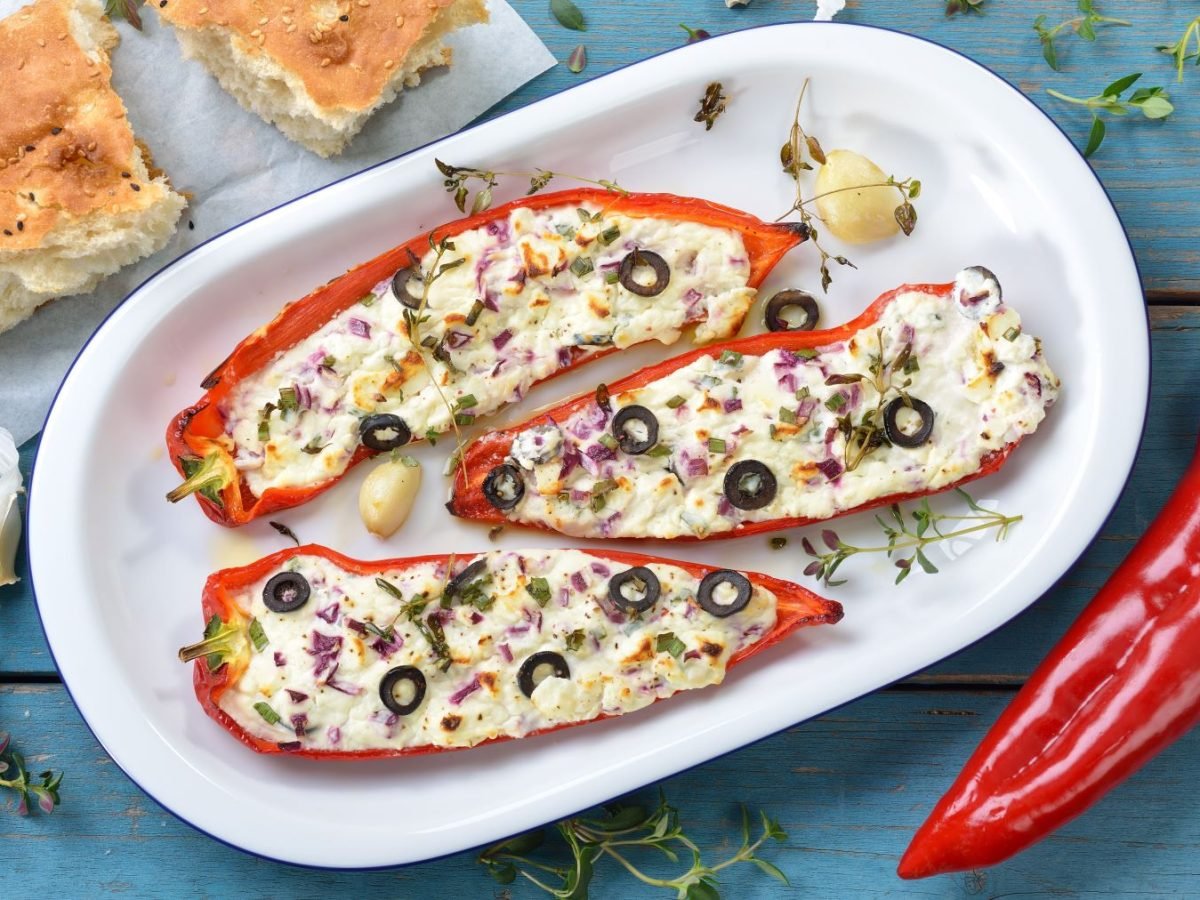 Paprika mit Feta-Füllung auf einem weißen Teller vor blauem Hintergrund. Auf dem Hintergrund Fladenbrot, Paprika, Thymian und Knoblauch verteilt.