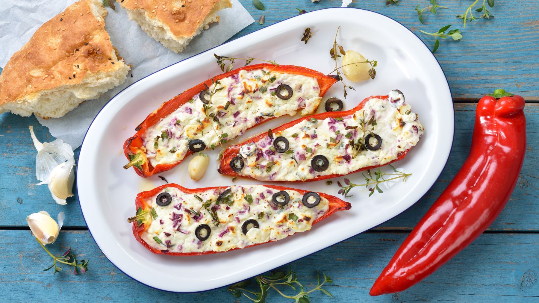 Paprika mit Feta-Füllung auf einem weißen Teller vor blauem Hintergrund. Auf dem Hintergrund Fladenbrot, Paprika, Thymian und Knoblauch verteilt.