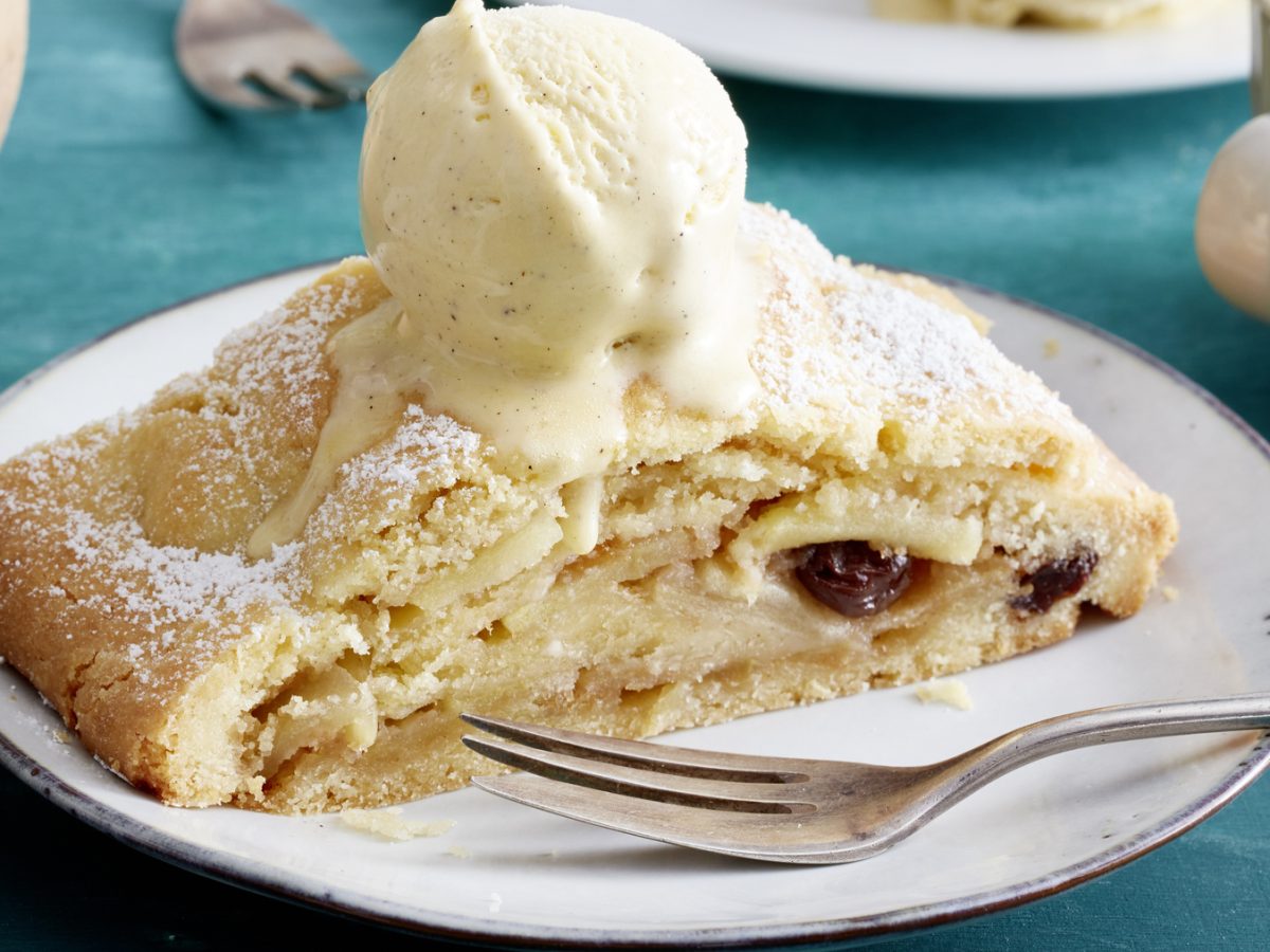 Ein Stück Südtiroler Apfelstrudel auf einem Teller mit Gabel und einer Kugel Vanilleeis oben drauf.