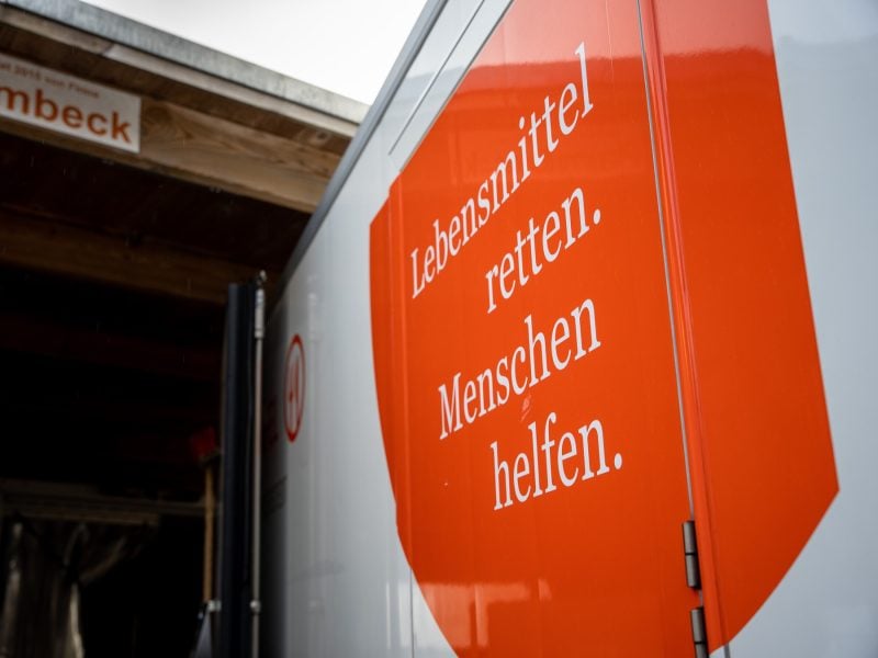 Weißer Lieferwagen von der Tafel, mit großem orangefarbenem Schild, darauf der Schriftzug „Lebensmittel retten. Menschen helfen.“, vor einem Holzunterstand.