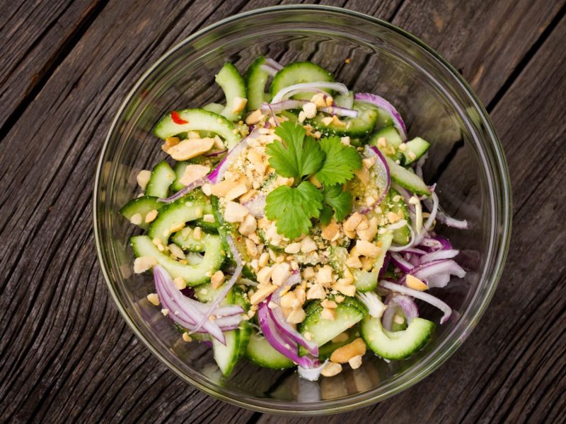 Frischer Thai-Gurkensalat mit roten Zwiebeln, gehackten Erdnüssen und Koriander in einer Glasschale auf rustikalem Holztisch.