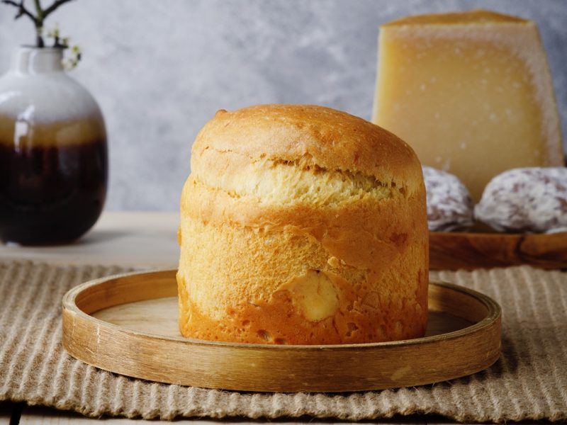 Goldbraunes, luftig gebackene Torta di Pasqua auf einem runden Holztablett, im Hintergrund ein Käseblock, Wurstscheiben und eine Vase.