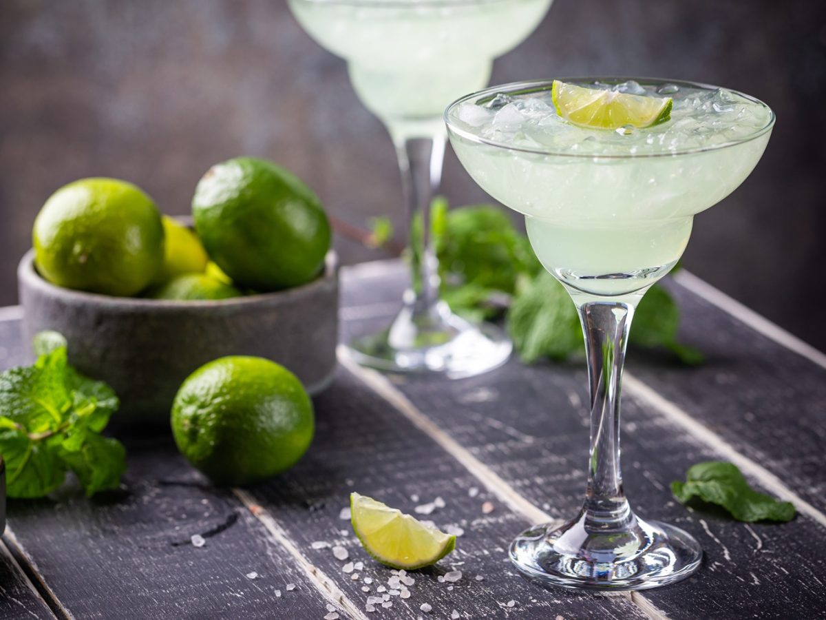 Glas mit einem Virgin Margarita, dass mit einem Limettenschnitz dekoriert ist. Im Hintegrrund ein weiterer Mocktail und eine Schale mit frischen Limetten.
