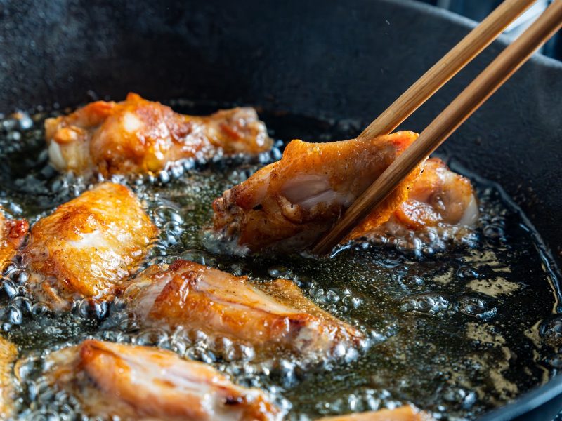Frittierte Hähnchenflügel, die gerade mit zwei Essstäbchen beim Frittieren gewendet werden. Alles in Nahaufnahme von der Seite.