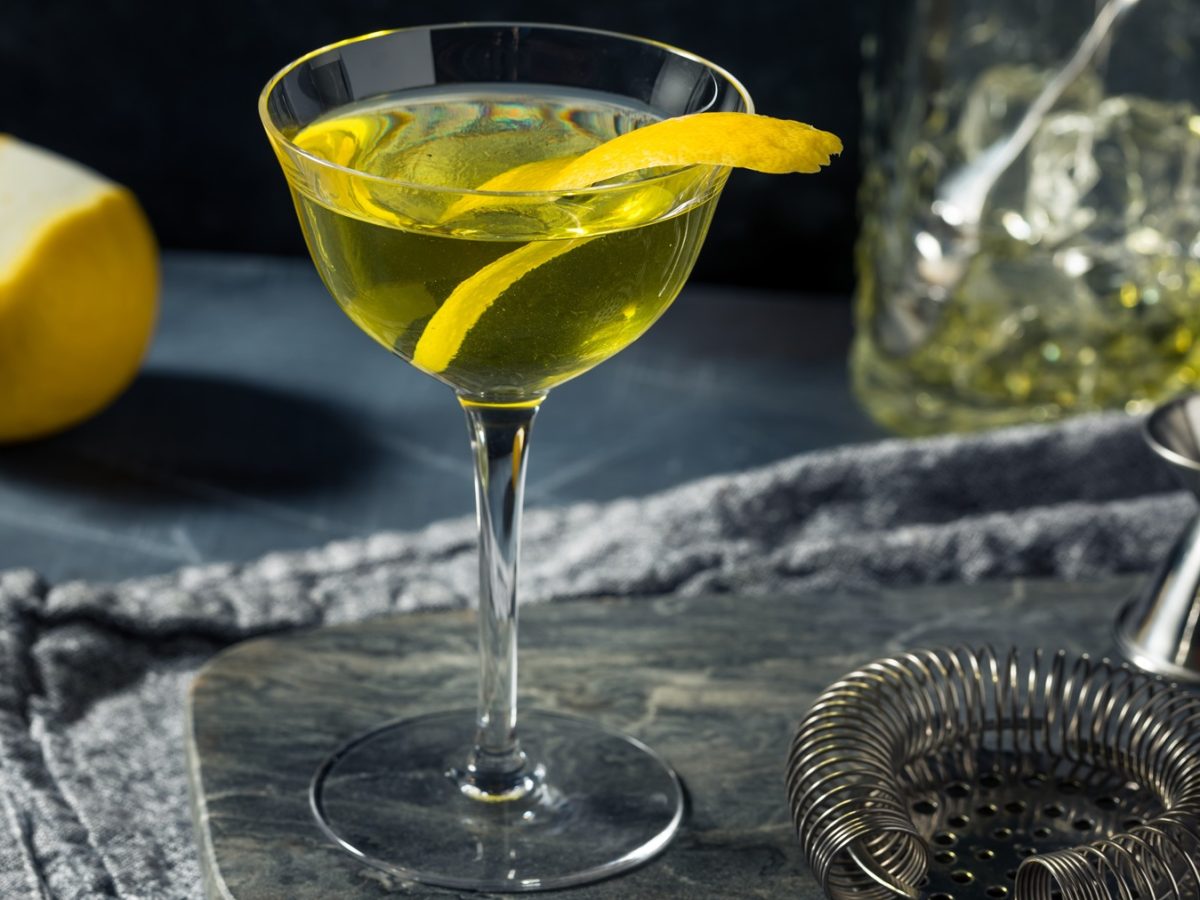 Ein klassischer Alaska-Cocktail in einem hohen Glas mit geschwungener Zitronenschale, daneben eine Zitrone, Barsieb und Messbecher.