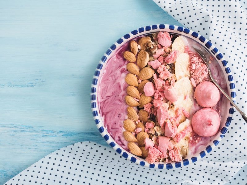 Schale mit Beeren-Joghurtbowl, Mandeln, Bananenscheiben, pinken Fruchtdrops von Sunday Natural und Pralinen auf hellblauem Holztisch mit gepunktetem Tuch.