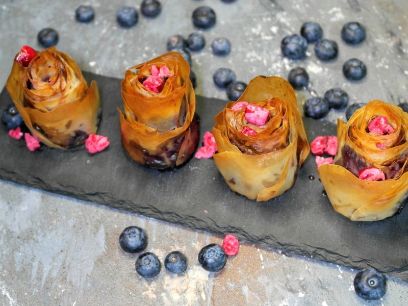 Vier Blaubeer-Rosen auf einer schwarzen länglichen Schiefertafel und grauem Steintisch mit Blaubeeren und grauer Decke. Alles von schräg oben fotografiert.