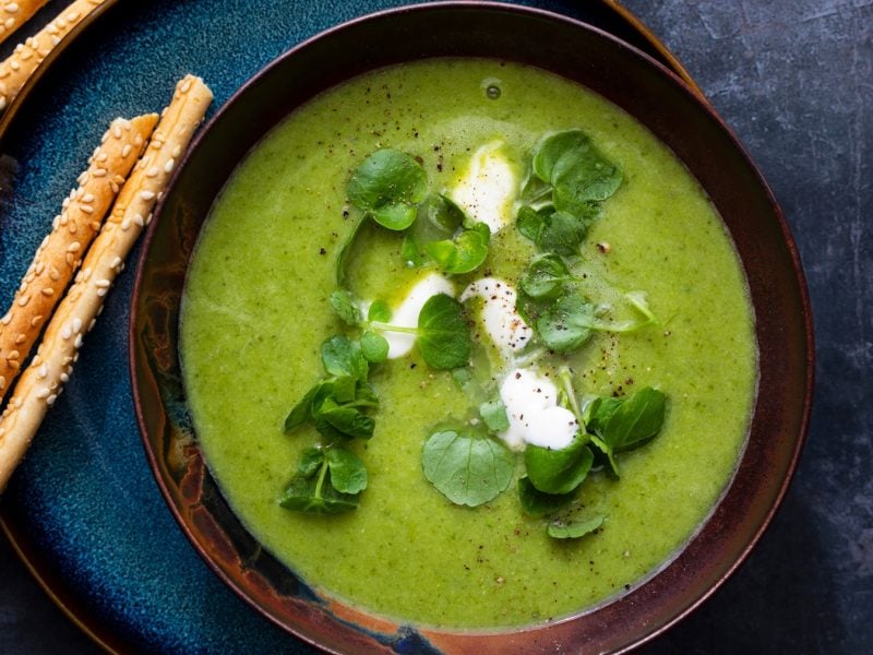 Eine dunkelblaue Schale mit der Brunnenkresse-Suppe auf einem dunklen Tisch. Daneben noch ein paar Gebäckstangen. Alles direkt von oben fotografiert.