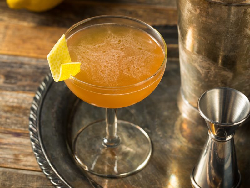 Ein eleganter Champs-Élysées-Cocktail mit bernsteinfarbener Flüssigkeit und Zitronengarnitur auf einer rustikalen Holzoberfläche.