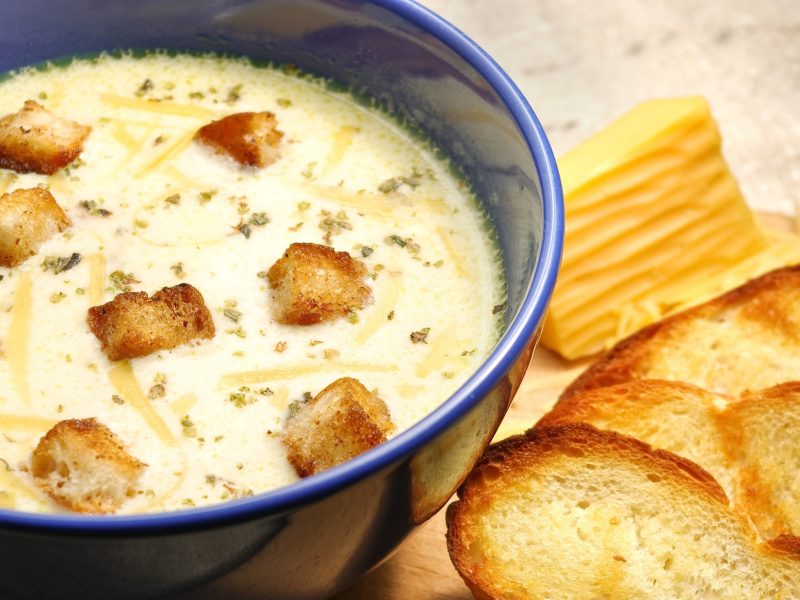 Draufsicht: Eine Käsesuppe in einer runden, dunklen Suppenschüssel. Daneben liegen geröstete Brotscheiben.