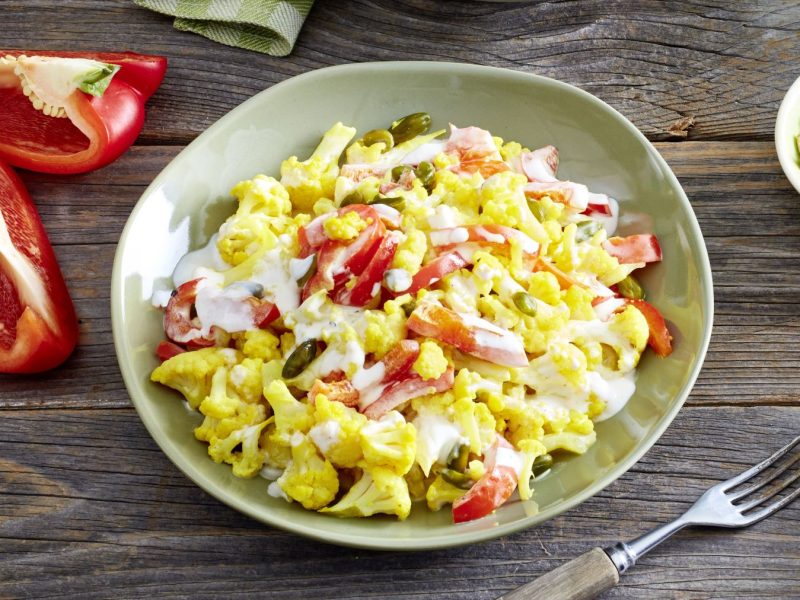 Blumenkohl-Paprika-Salat in einer grünen Schüssel auf hölzernem Hintergrund. Links Paprika und rechts auf dem Foto Pinienkerne.