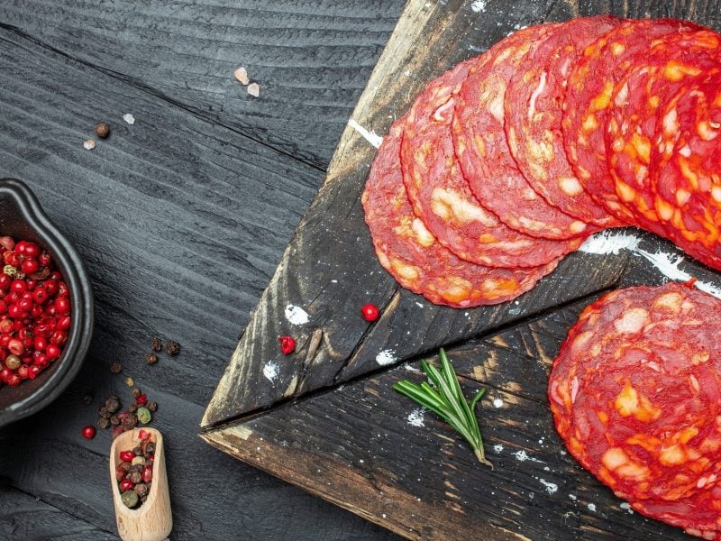 Draufsicht: mehrere Scheiben Chorizo auf einer Platte aus Schiefer. Die liegt auf einem Untergrund aus Holz. Daneben ist eine Schale mit Pfefferkörnern.