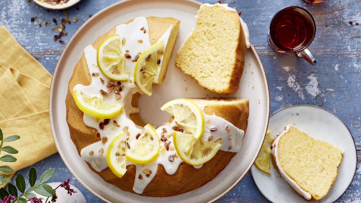 Earl-Grey-Zitronen-Gugelhupf auf einem weißen Teller. Auf dem Hintergrund Tee, ein Stück Kuchen auf einem kleinen Teller, ein gelbes Geschirrtuch und ein Blumenstrauß verteilt.