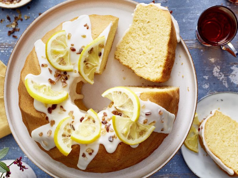 Earl-Grey-Zitronen-Gugelhupf auf einem weißen Teller. Auf dem Hintergrund Tee, ein Stück Kuchen auf einem kleinen Teller, ein gelbes Geschirrtuch und ein Blumenstrauß verteilt.