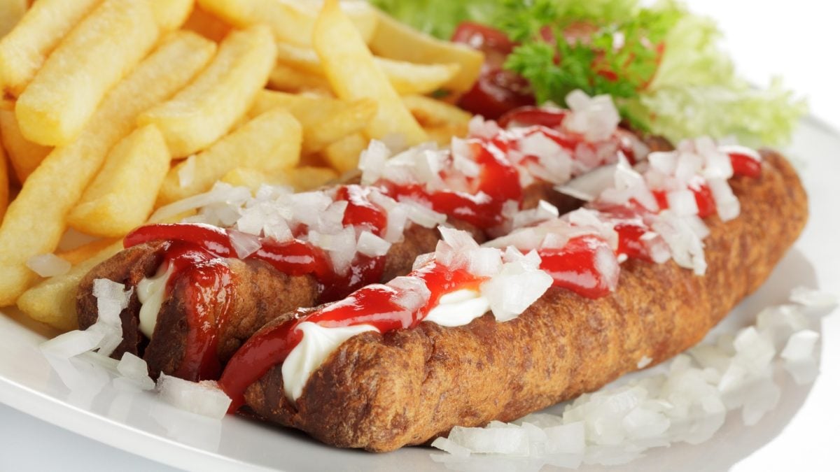 Frontal: Zwei frittierte Würste mit Pommes auf einem Teller. Daneben sind Mayonnaise und Ketchup.