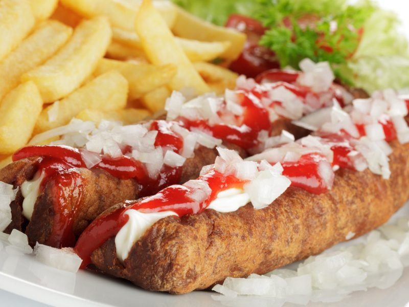 Frontal: Zwei frittierte Würste mit Pommes auf einem Teller. Daneben sind Mayonnaise und Ketchup.