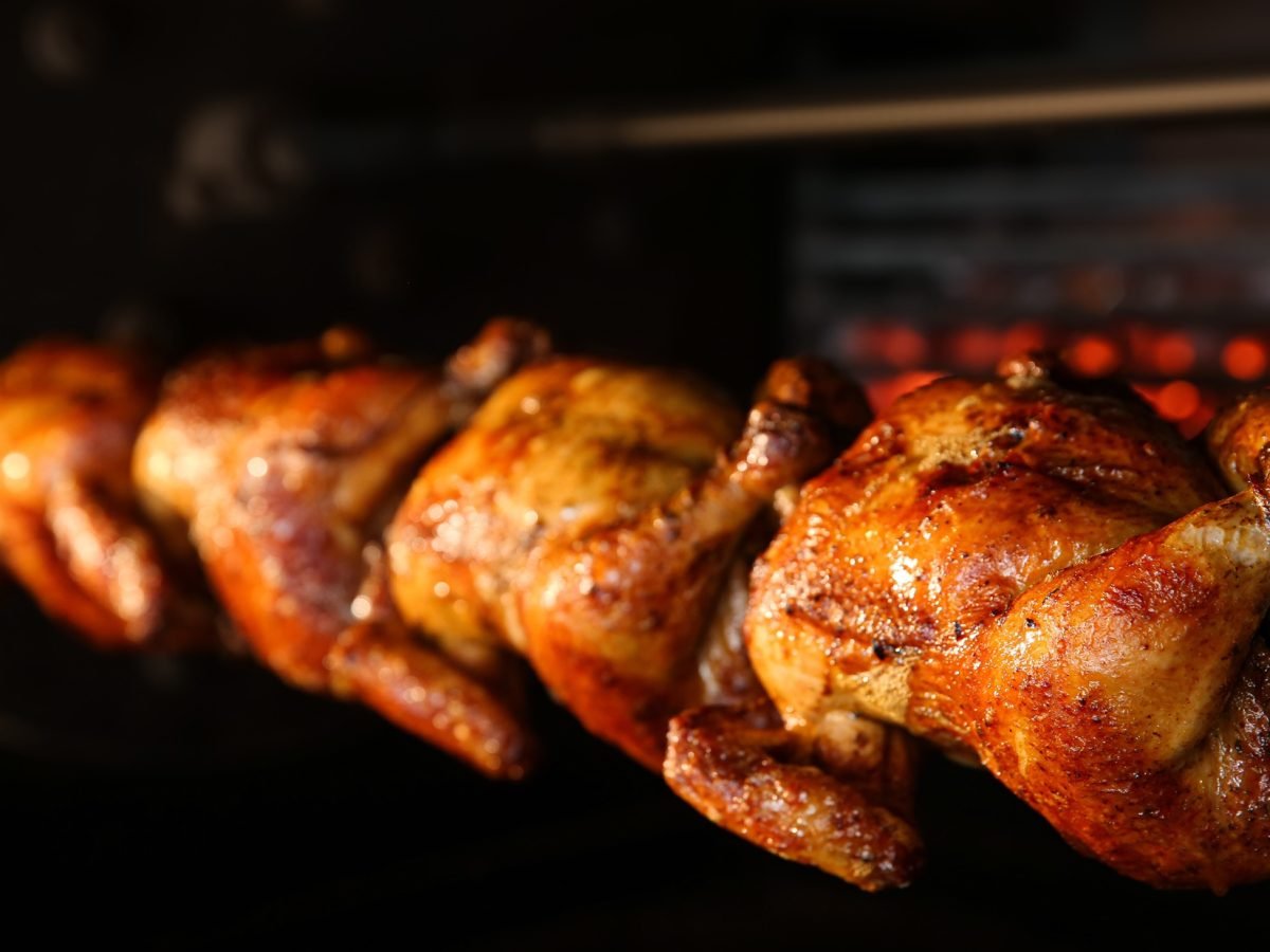 Frontal: Vier Grillhähnchen auf einem Spieß in einem Ofen.
