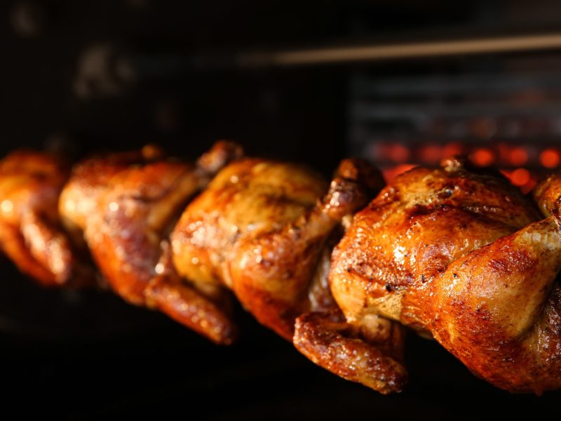 Frontal: Vier Grillhähnchen auf einem Spieß in einem Ofen.