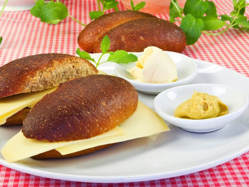 Zwei Käsebrötchen auf einem hellen Teller. Im Hintergrund ist ein Brötchen ohne Käse. Auf dem Teller ist eine kleine Schale mit Senf und eine Mit Butter.