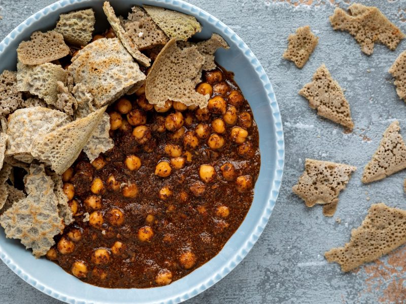 Draufsicht: Auf hellem Grund aus Holz steht eine runde Schale mit gekochten Kichererbsen und kleinen gezupften Brotstücken, Weitere kleine Brotfetzen liegen rechts neben der Schüssel.