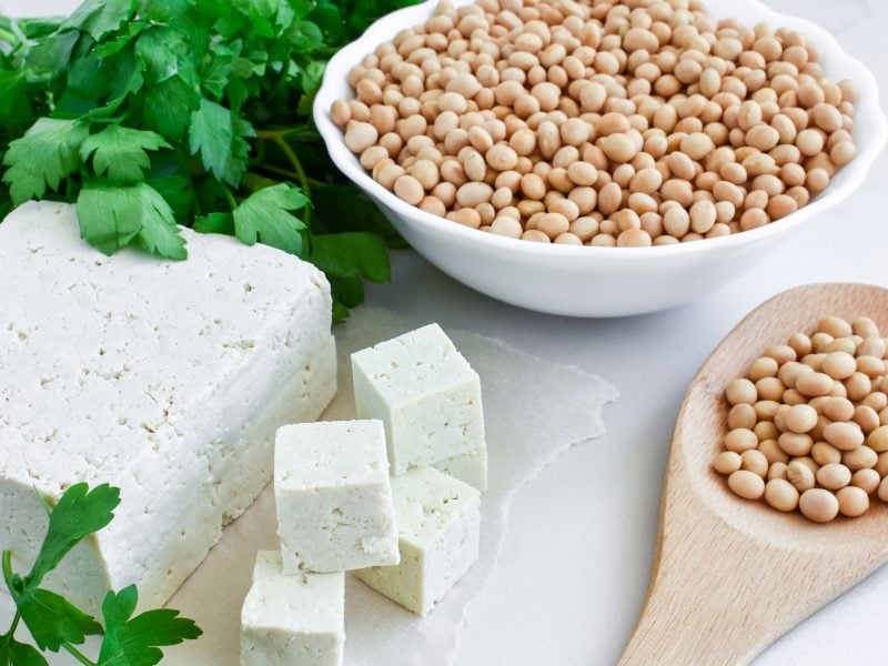 Draufsicht, schräg: Ein heller Untergrund, auf dem eine großer Block und einige kleine Würfel Tofu liegen. Und den Tipp liege verteilt frische Petersilie. Daneben ist eine rund weiße Schüssel und ein Holzlöffel mit rohen Sojabohnen.