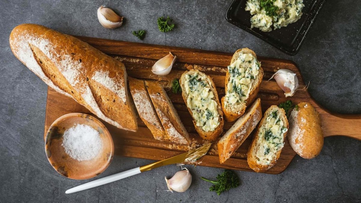 Draufsicht: Dunkler Untergrund, ein Holzbrett, auf dem eine längliches Brot liegt. Das ist teilweise angeschnitten. Auf den Scheiben ist eine kräutercreme. Daneben liegen ein Messer und Knoblauchzehen.. Auch ein rundes Gefäß mit groben Salzkristallen ist daneben.