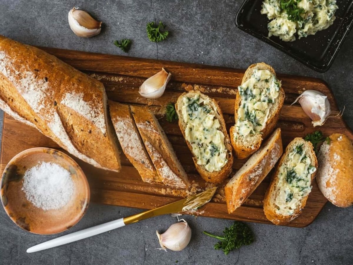 Draufsicht: Dunkler Untergrund, ein Holzbrett, auf dem eine längliches Brot liegt. Das ist teilweise angeschnitten. Auf den Scheiben ist eine kräutercreme. Daneben liegen ein Messer und Knoblauchzehen.. Auch ein rundes Gefäß mit groben Salzkristallen ist daneben.