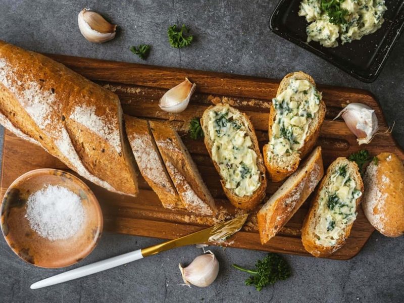 Draufsicht: Dunkler Untergrund, ein Holzbrett, auf dem eine längliches Brot liegt. Das ist teilweise angeschnitten. Auf den Scheiben ist eine kräutercreme. Daneben liegen ein Messer und Knoblauchzehen.. Auch ein rundes Gefäß mit groben Salzkristallen ist daneben.
