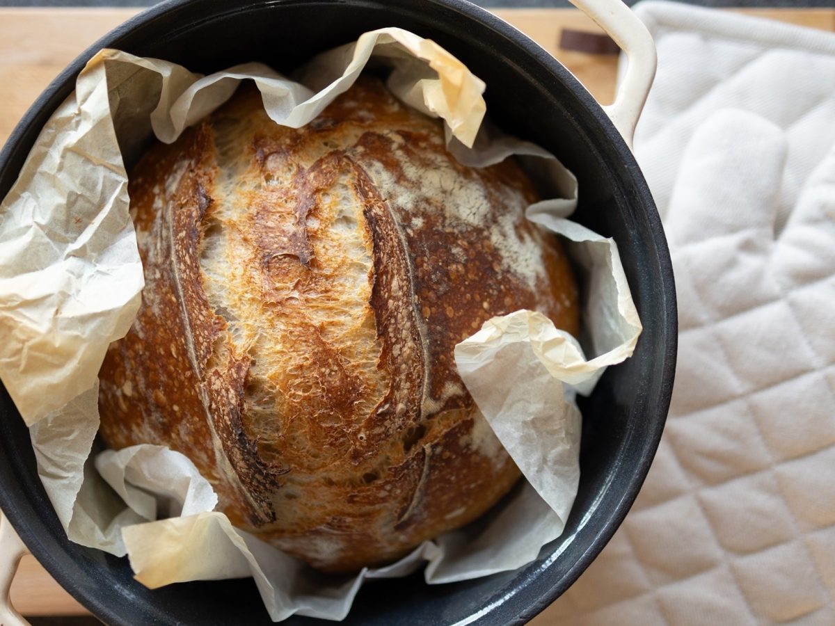 Draufsicht: Ein rundes gebackenes Brot in einem runden, offenen Topf. Der steht auf einem Holzbrett. Daneben liegt ein Topflappen.