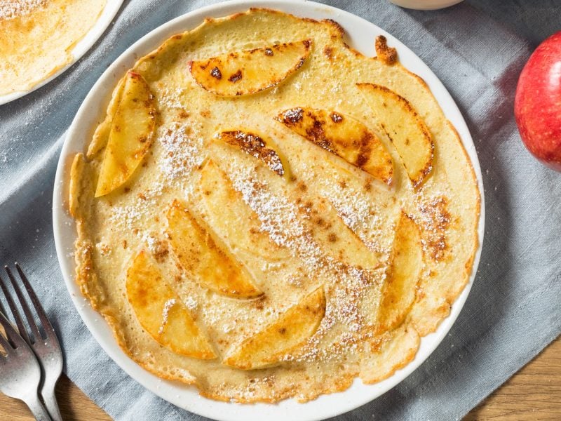 Draufsicht: Ein runder Pfannkuchen mit Apfel auf einem runden Teller. Neben dem liegt ein frischer Apfel.