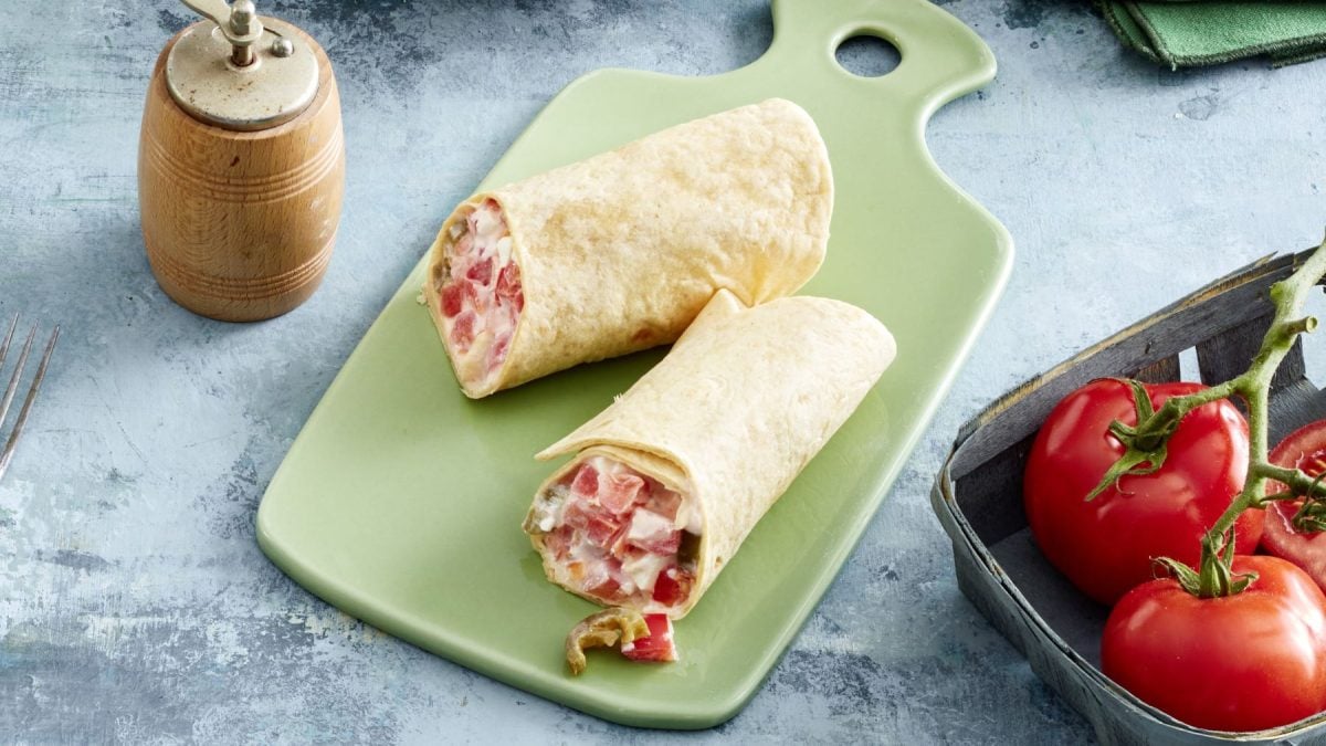 Paprika-Wrap auf einem grünen Schneidebrett auf grauem Hintergrund. Links eine Pfeffermühle, rechts Tomaten.