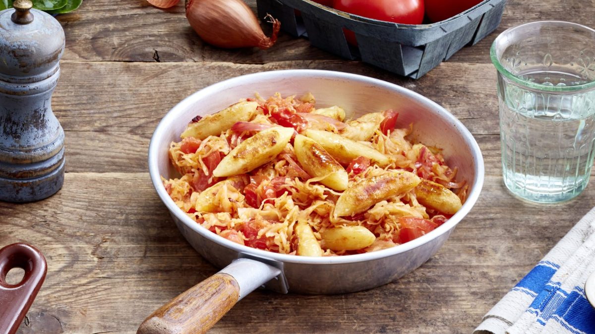 Schupfnudel-Sauerkraut-Pfanne auf hölzernem Hintergrund. Rechts ein Glas Wasser und links eine Pfeffermühle.