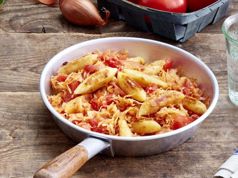 Schupfnudel-Sauerkraut-Pfanne auf hölzernem Hintergrund. Rechts ein Glas Wasser und links eine Pfeffermühle.
