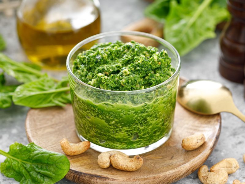 Draufsicht, leicht schräg: Ein offener Behälter aus Glas mit einem Spinat-Pesto mit Cashews. Drum herum liegen oder stehen u.a. frische Spinatblätter, Cashewkerne, eine Karaffe mit Öl, eine Suppenlöffel und zwei Gewürzmühlen aus Holz.