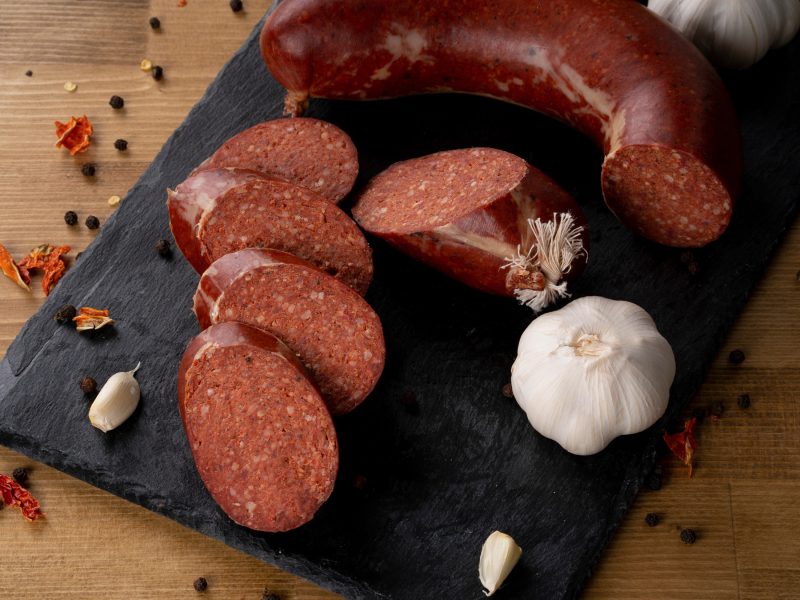 Draufsicht: Eine angeschnittene Wurst sowie einige Wurstscheiben auf einem Brett aus Schiefer.