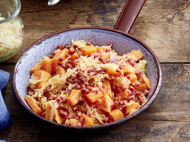 Süßkartoffel-Sauerkraut-Pfanne in einer Pfanne auf hölzernem Hintergrund. Links davon ein blaues Geschirrtuch.