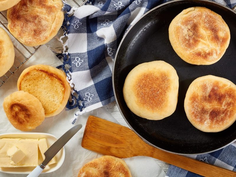 Draufblick: mehrere Toasties aus der Pfanne liegen auf einem hellen Untergrund. Einige English Muffins sind in einer dunklen Pfanne, die anderen liegen daneben.