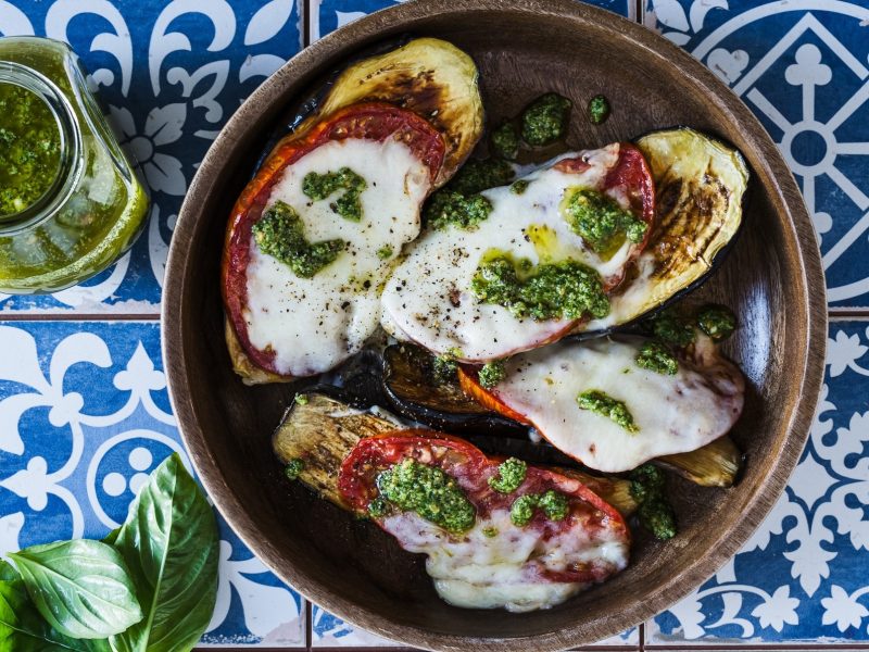 Draufsicht: Ein runder Teller. Auf der liegen vier mit Mozzarella überbackenen Scheiben Aubergine. Daneben liegen frische Basilikumblätter und eine kleines Glas mit einem Kräuteröl, dass an ein Basilikumpesto erinnert.