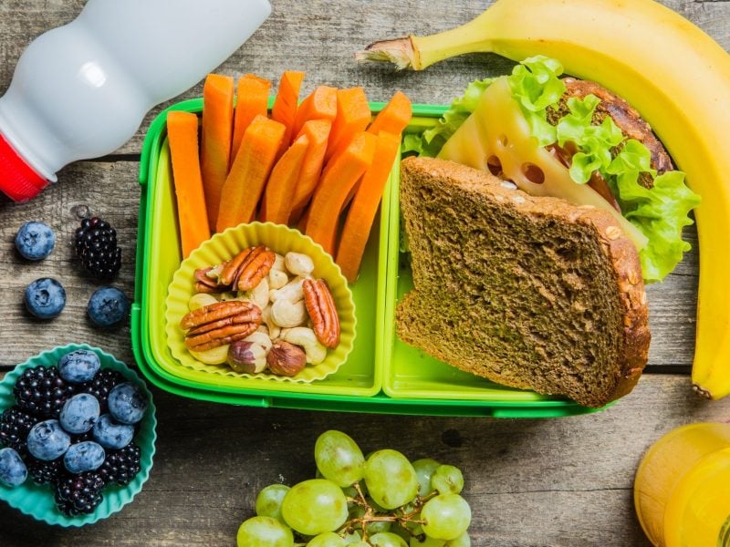 Draufsicht: Eine Brotdose aus Plastik, die mit Möhrensticks und einem Käsebrot befüllt ist. Drum herum liegen Weintrauben, Beeren, zwei Trinkflaschen aus Plastik und eine Banane.