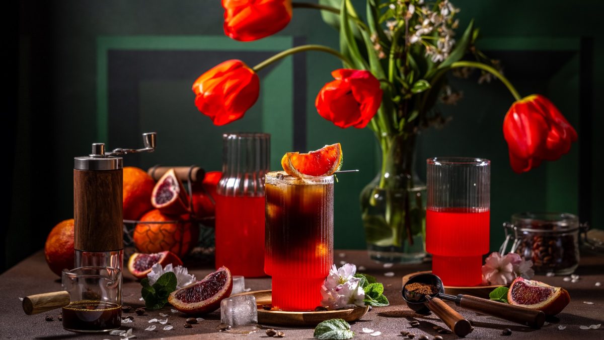 Stimmungsvolle Drink-Szene mit Espresso-Blutorangen-Tonic Blutorangen, Holzmühle und leuchtenden Tulpen vor dunkelgrünem Hintergrund.