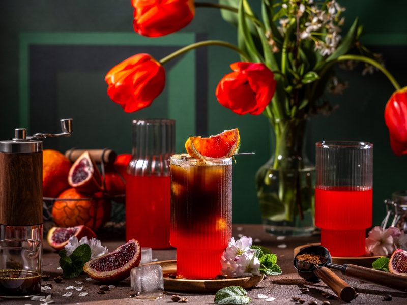 Stimmungsvolle Drink-Szene mit Espresso-Blutorangen-Tonic Blutorangen, Holzmühle und leuchtenden Tulpen vor dunkelgrünem Hintergrund.