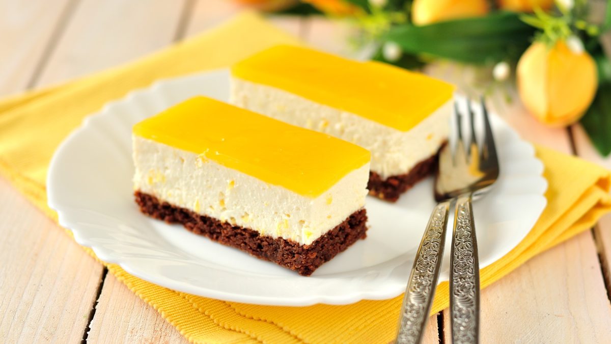 Zwei Stücke vom Fantakuchen auf einem weißen Teller und gelber Serviette. Alles auf einem Holztisch mit gelben Tulpen im Hintergrund.