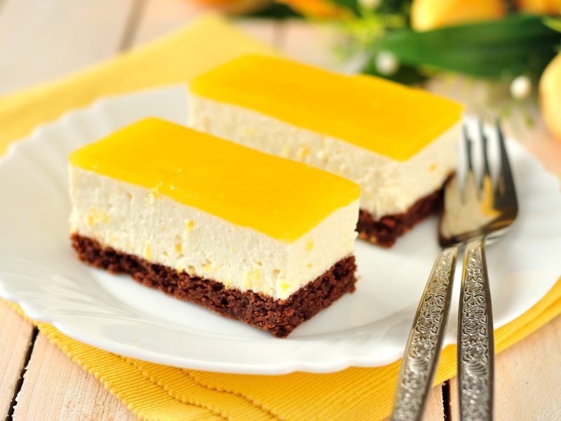 Zwei Stücke vom Fantakuchen auf einem weißen Teller und gelber Serviette. Alles auf einem Holztisch mit gelben Tulpen im Hintergrund.